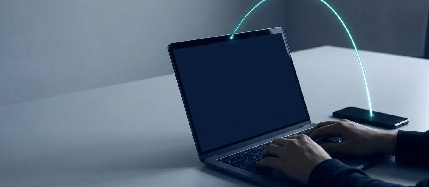 Manos sobre un portátil junto a un teléfono; una señal une ambos equipos